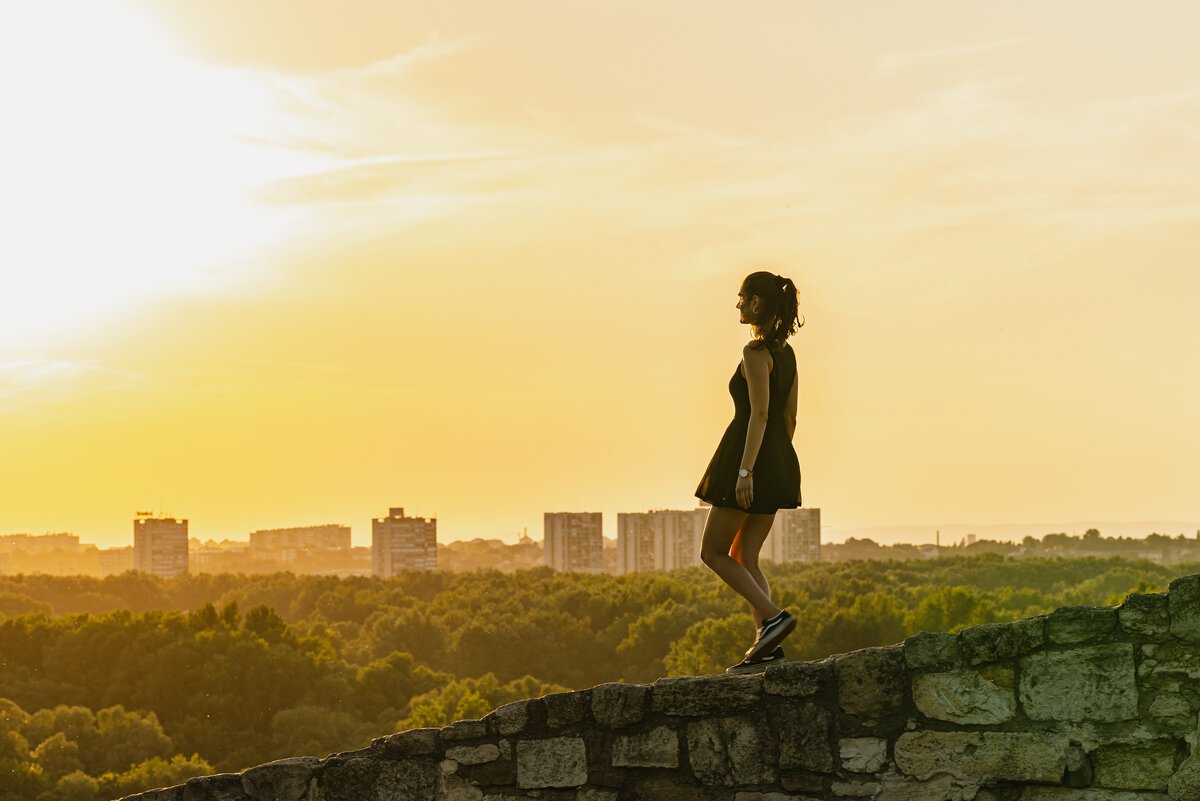 Woman in black dress silhouetted on a stone wall against golden sunset skyline