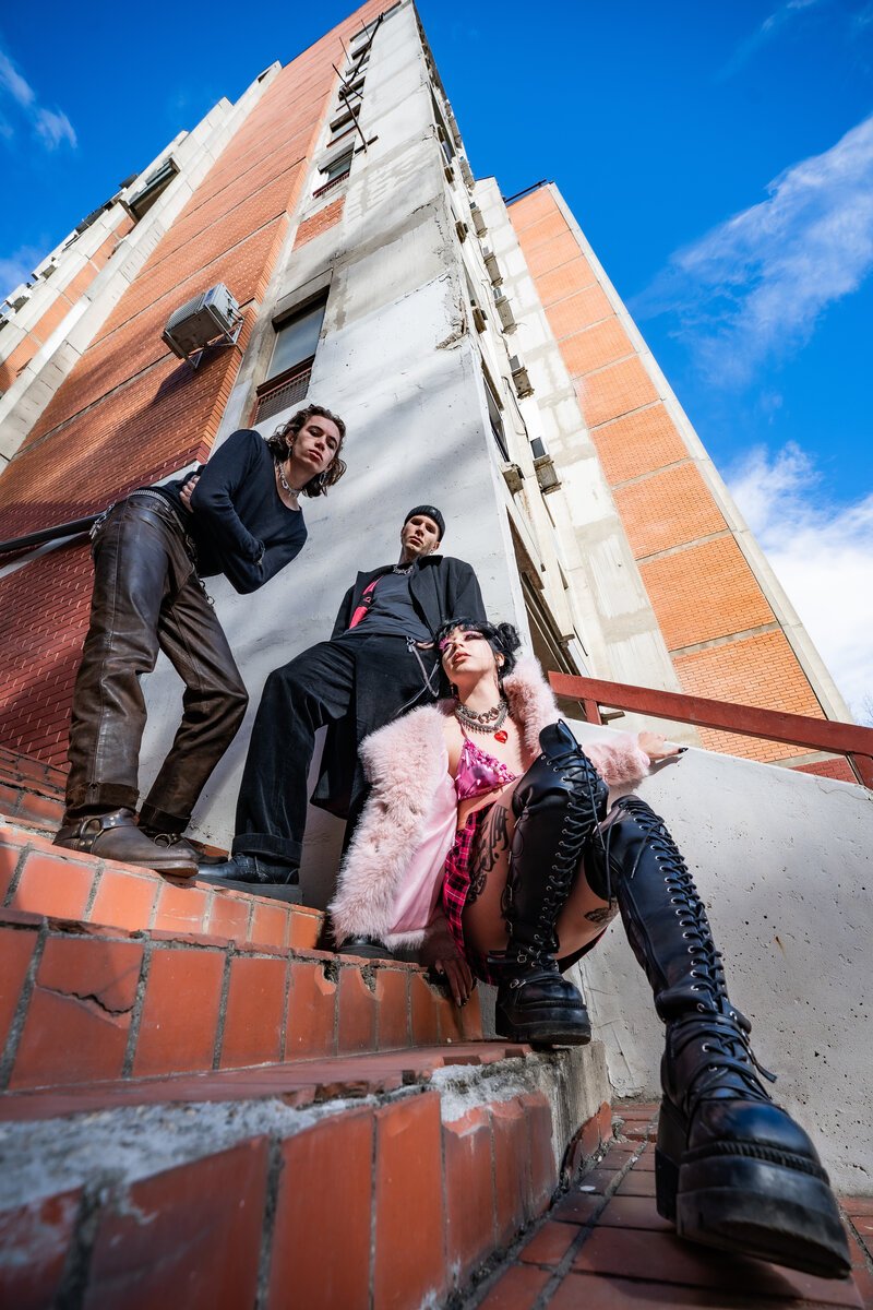Three styled subjects in edgy streetwear/glam looks (pink fur, corset, tall lace-up boots) posed low-angle on brick steps beneath a tower block