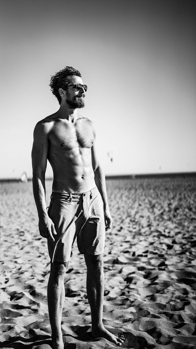Lean shirtless bearded man in sunglasses and shorts on a windswept beach, single-subject B&W portrait