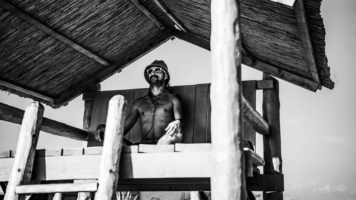 Shirtless man in beanie & sunglasses under a thatched beach hut