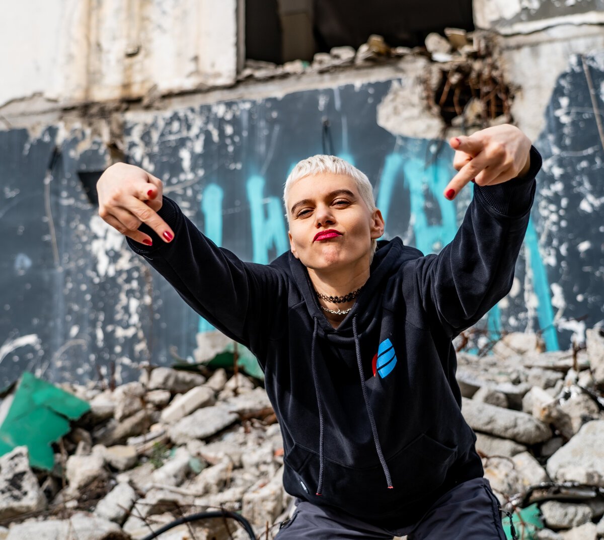 Woman in black branded hoodie pointing, crouched in rubble ruins