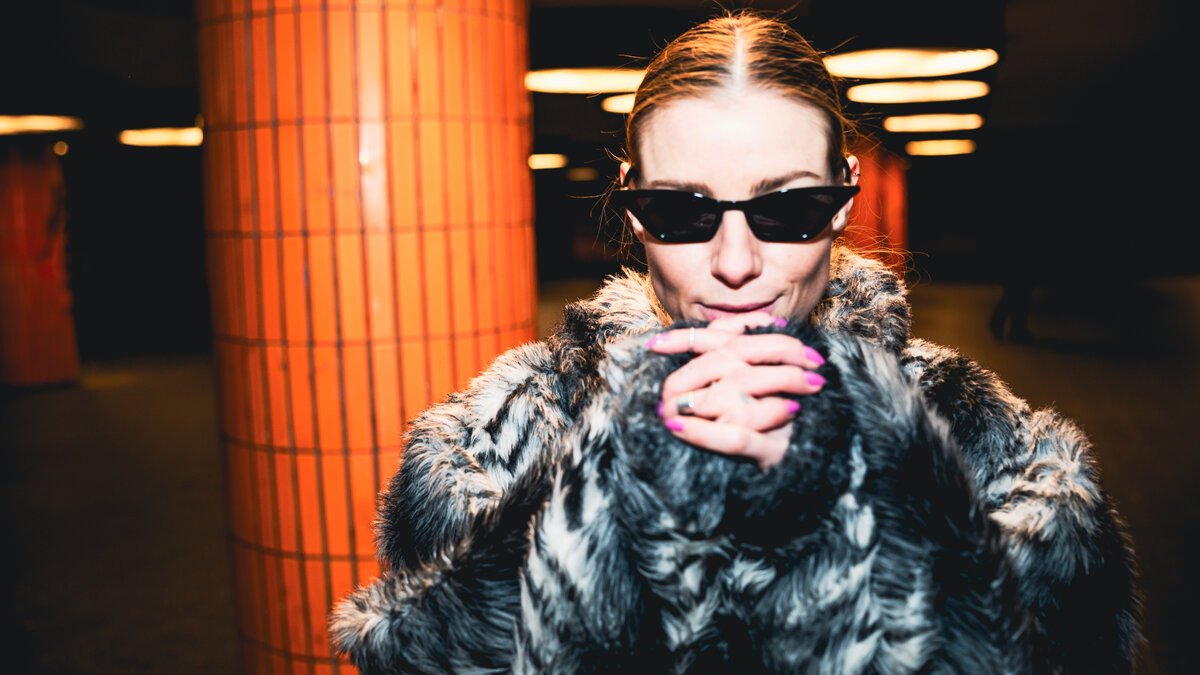 Woman in fur coat & sunglasses by orange-lit subway column at night