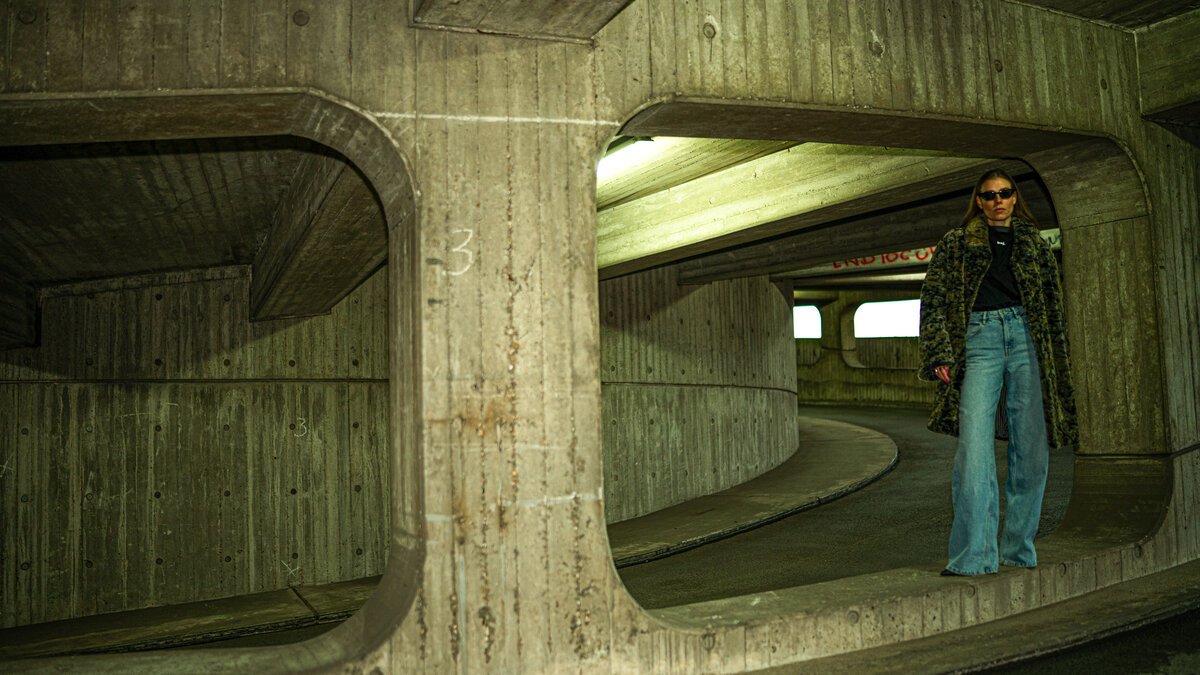 Woman in coat & wide jeans posed inside curved concrete parking structure