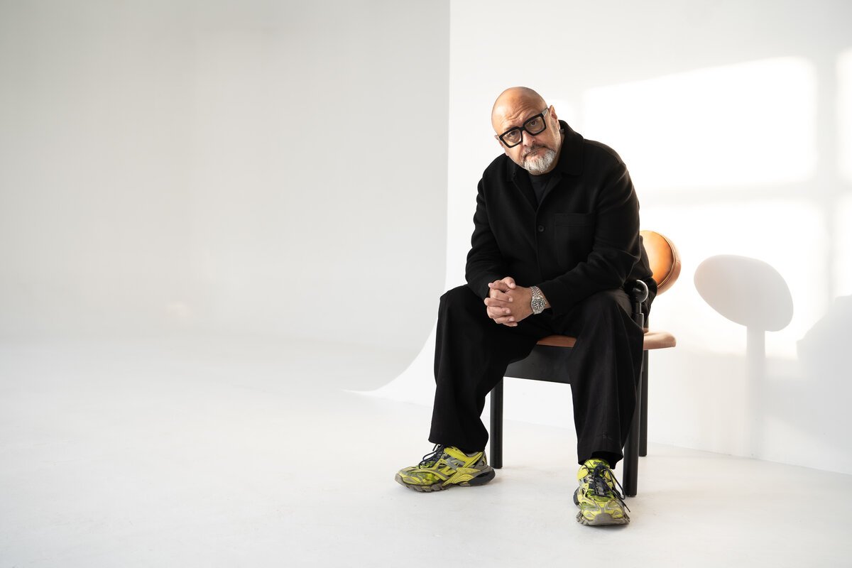 Bald bearded man in black seated on chair in bright white studio