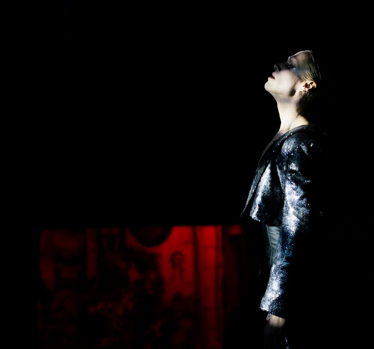 Profile of person in sequined jacket, head tilted up, red-lit religious iconography backdrop