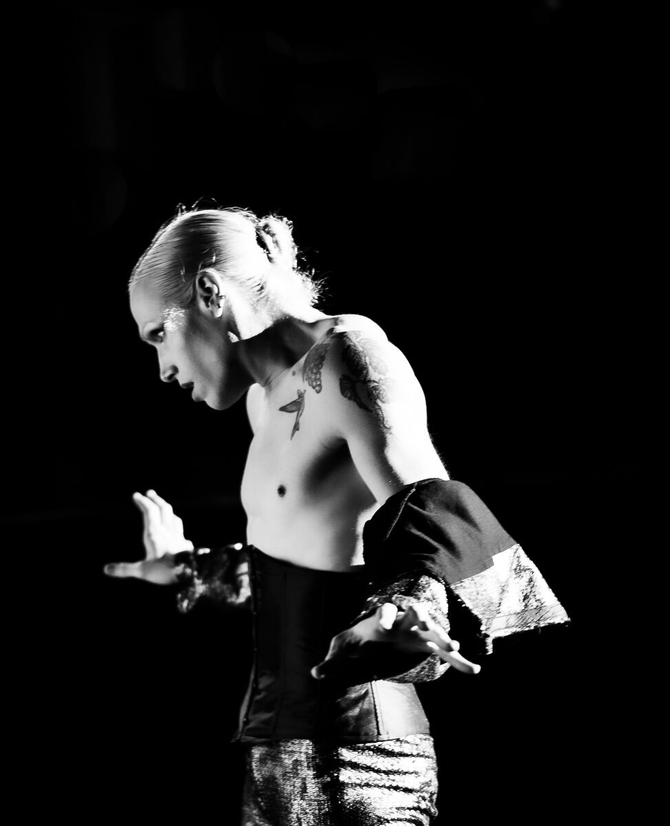 Shirtless tattooed performer with slicked hair, expressive hand gesture, against black