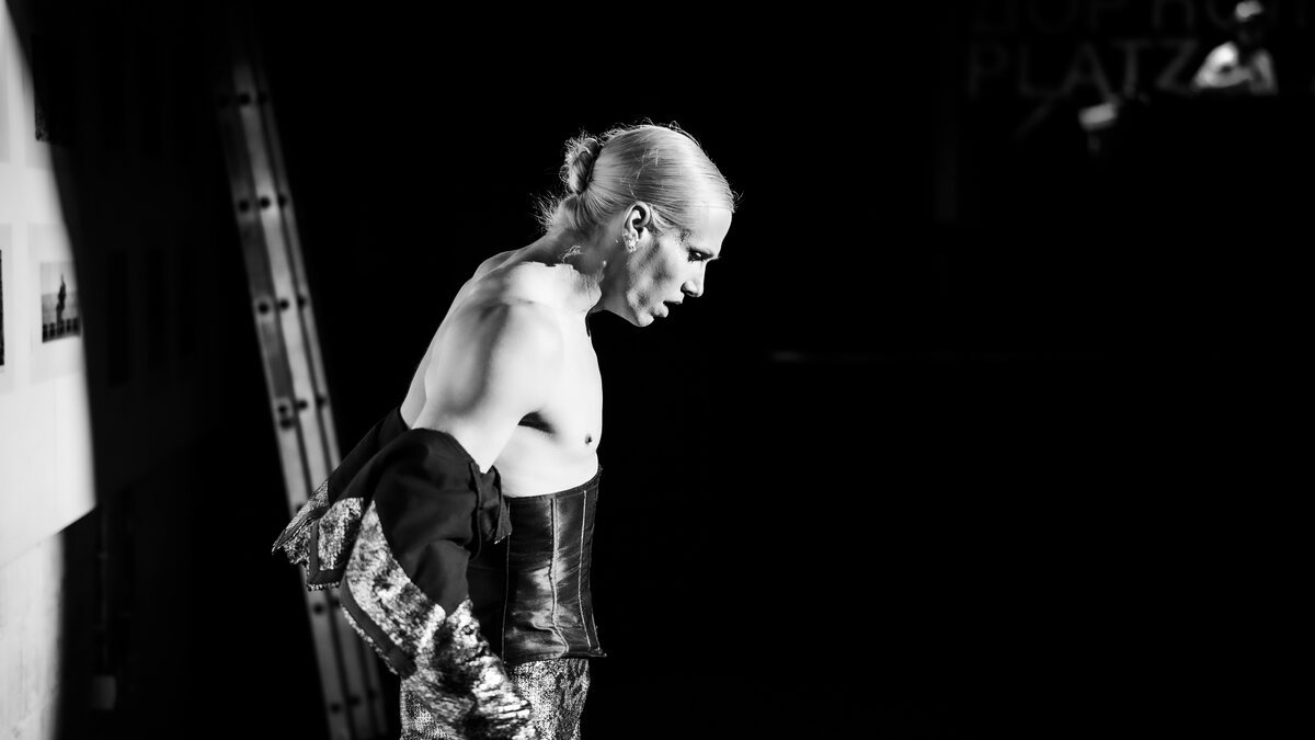 androgynous model backstage, slicked hair, leather/beaded look, bare torso