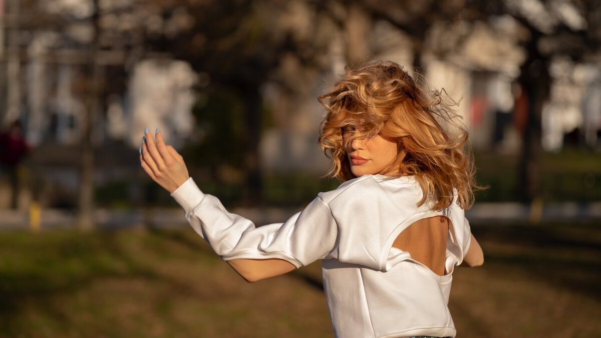 woman with windblown hair in white open-back top, golden-hour park