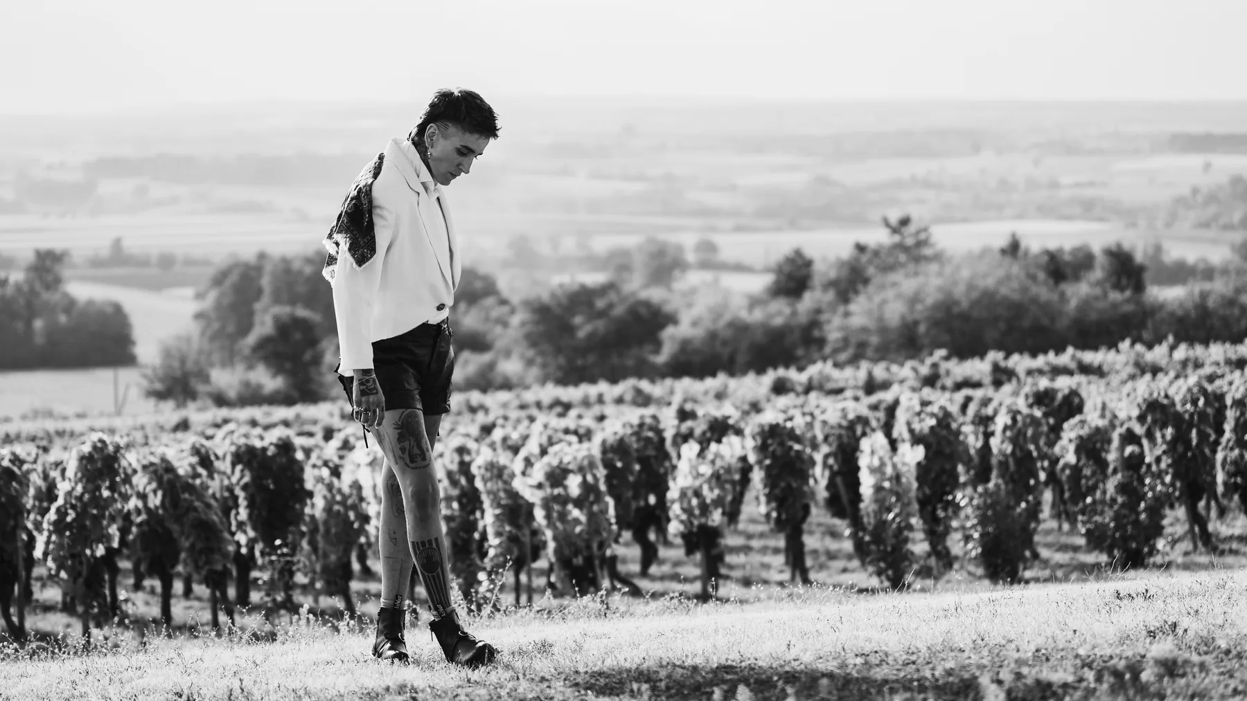 man in white jacket and shorts walking a vineyard hillside, tattoos