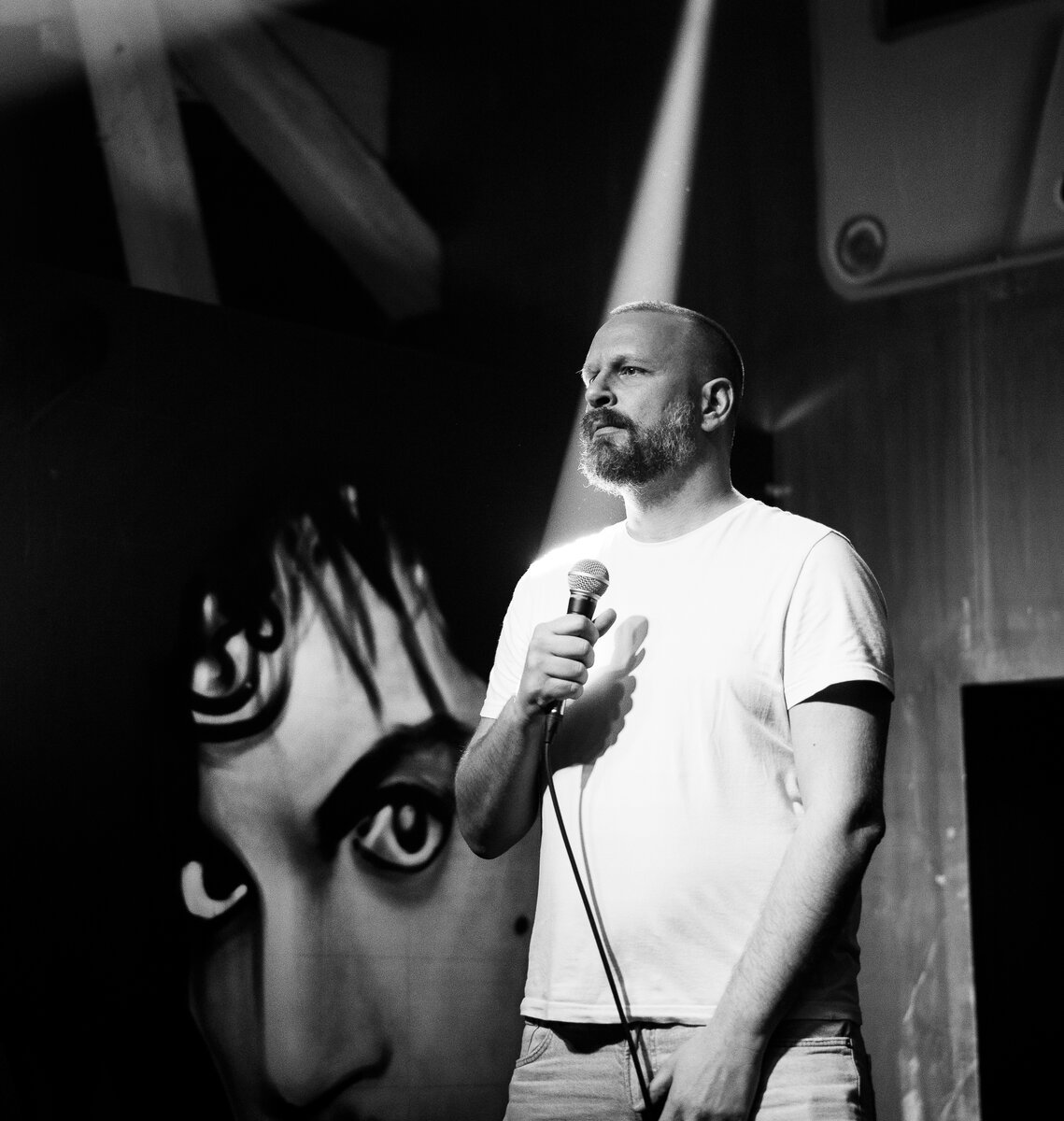 Bearded man holding microphone on stage beside a painted mural face