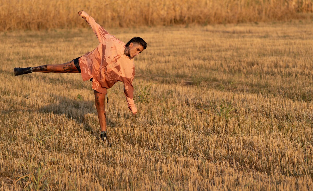 Man mid high-kick in a wheat field, golden hour, styled in pink jacket and shorts