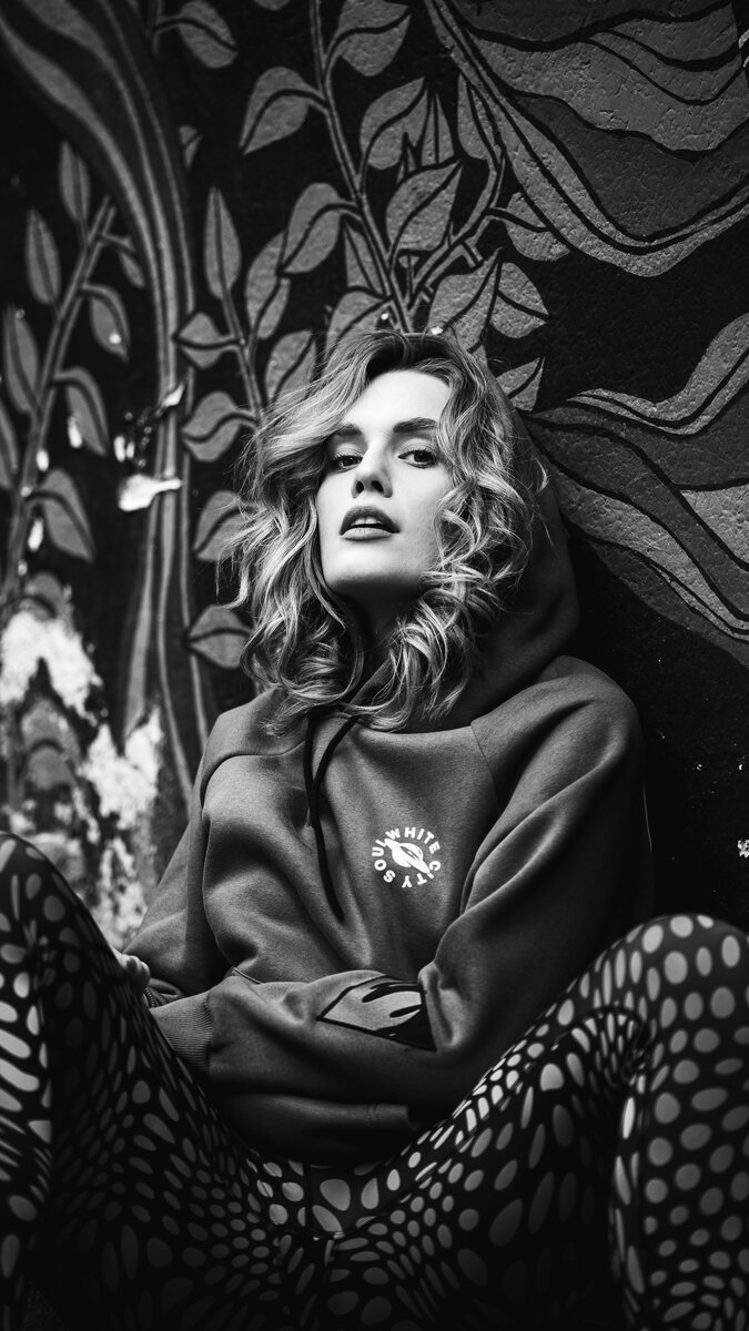 Blonde woman in hoodie and polka-dot tights seated against ornate patterned wall