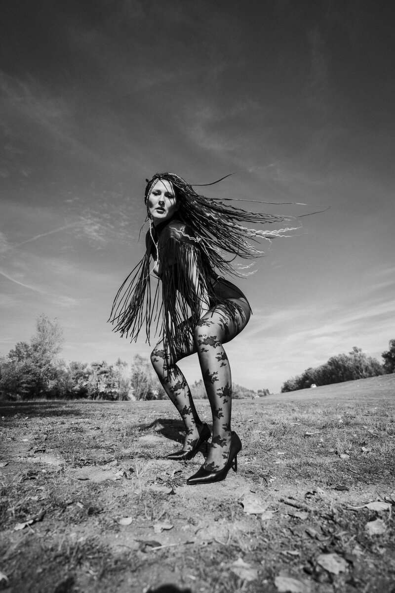 Woman with long braided fringe and heels posing dynamically in an open field, low angle against sky