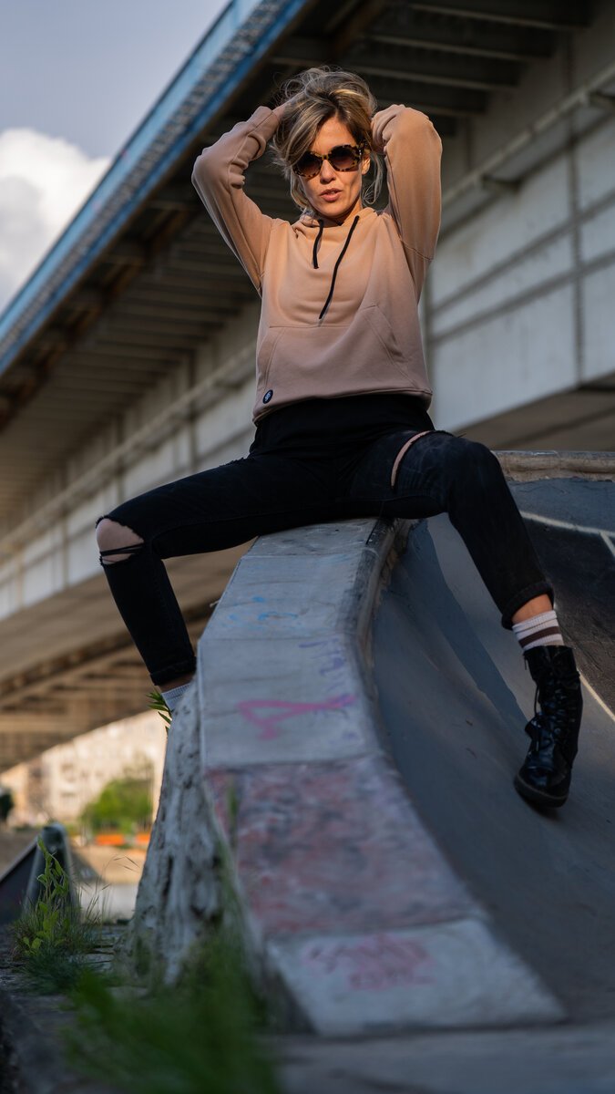 Woman in hoodie, sunglasses and ripped black jeans posing on a concrete skate ramp under a bridge