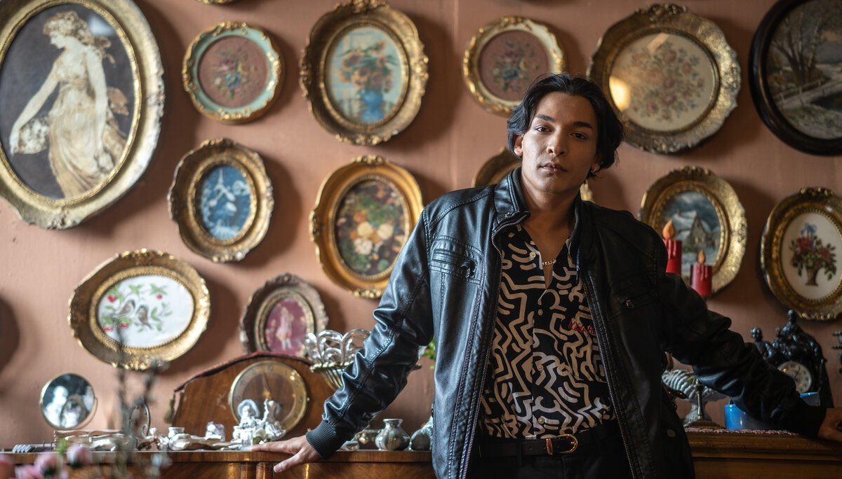 Young man in leather jacket and patterned shirt in an ornate room full of gilded oval frames