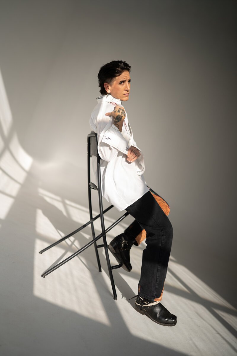 short-haired person perched on a metal stool in oversized white shirt, studio