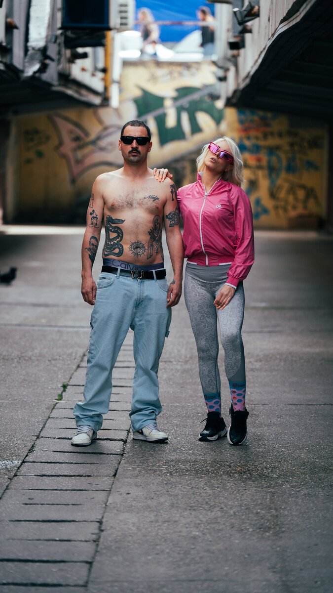 shirtless tattooed man and blonde woman in pink windbreaker, graffiti alley