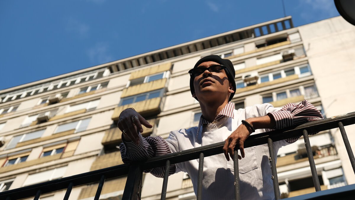 Young man on balcony railing, striped shirt, low angle against apartment block and blue sky