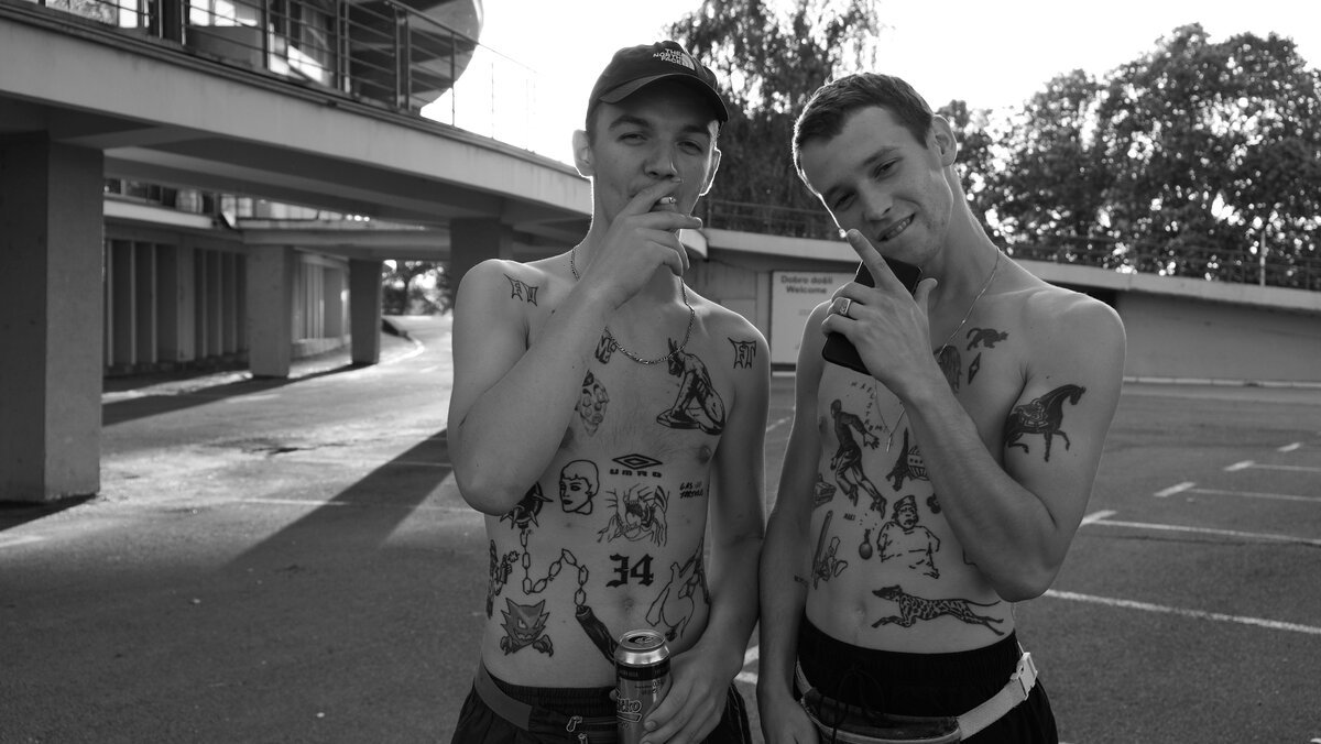 Two shirtless tattooed young men posing in a parking lot, one with beer