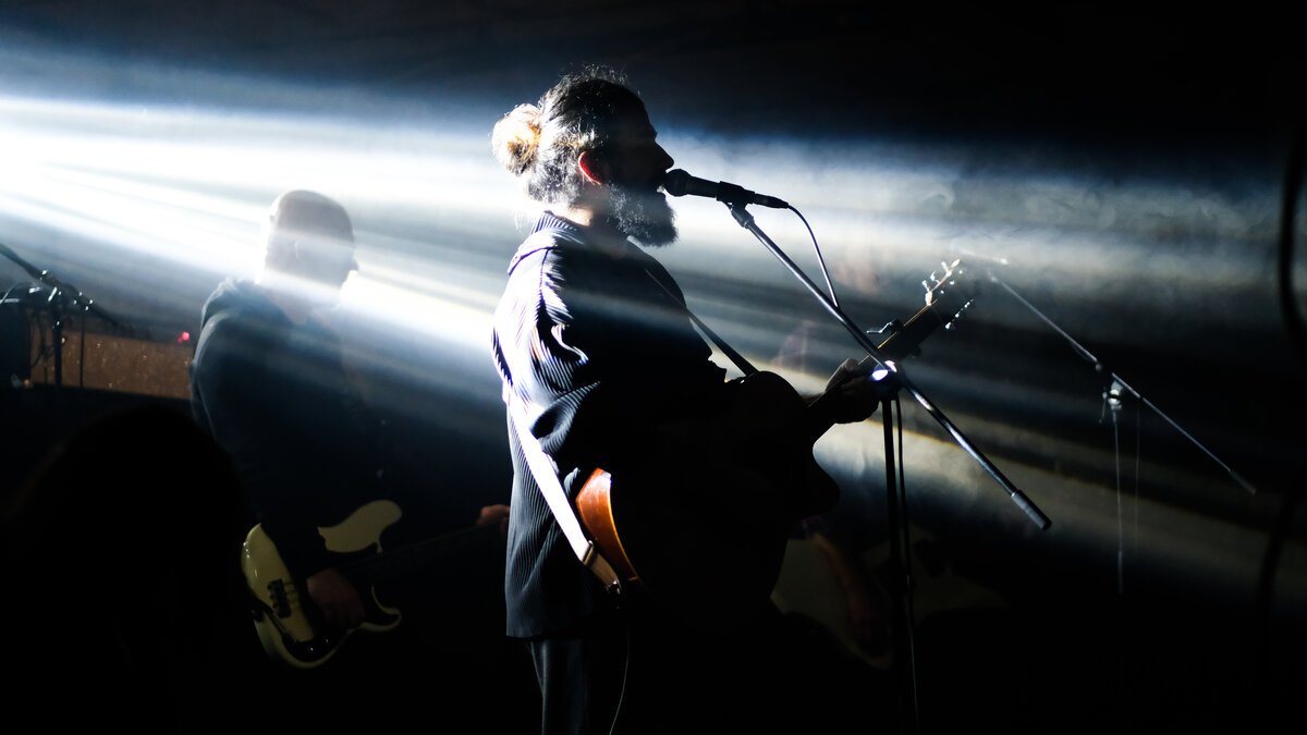 Bearded guitarist singing at mic on stage, backlit white beams, bassist behind