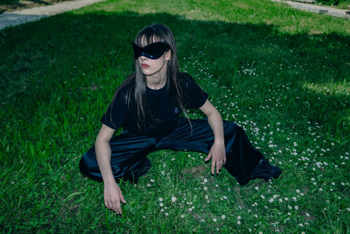 model in all-black streetwear crouching on grass, blindfold/mask