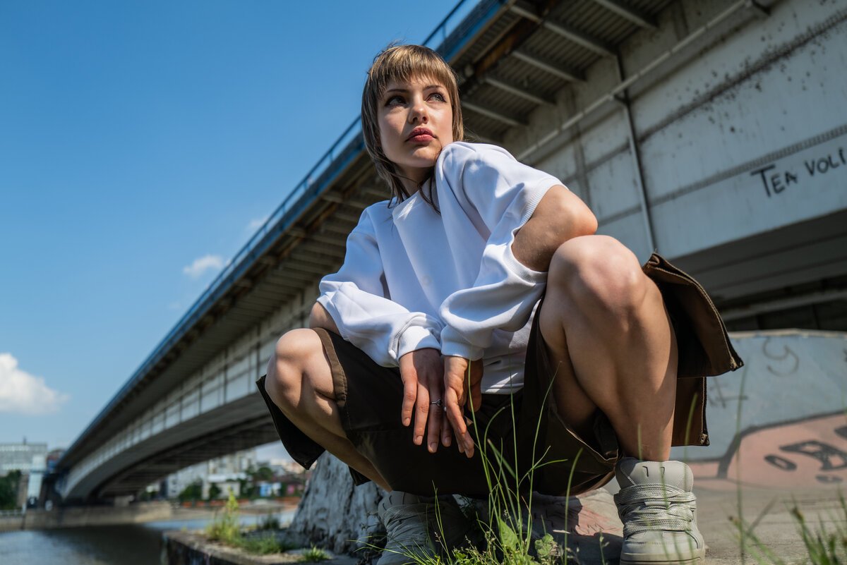 model crouching by riverside under bridge, white top, chunky sneakers