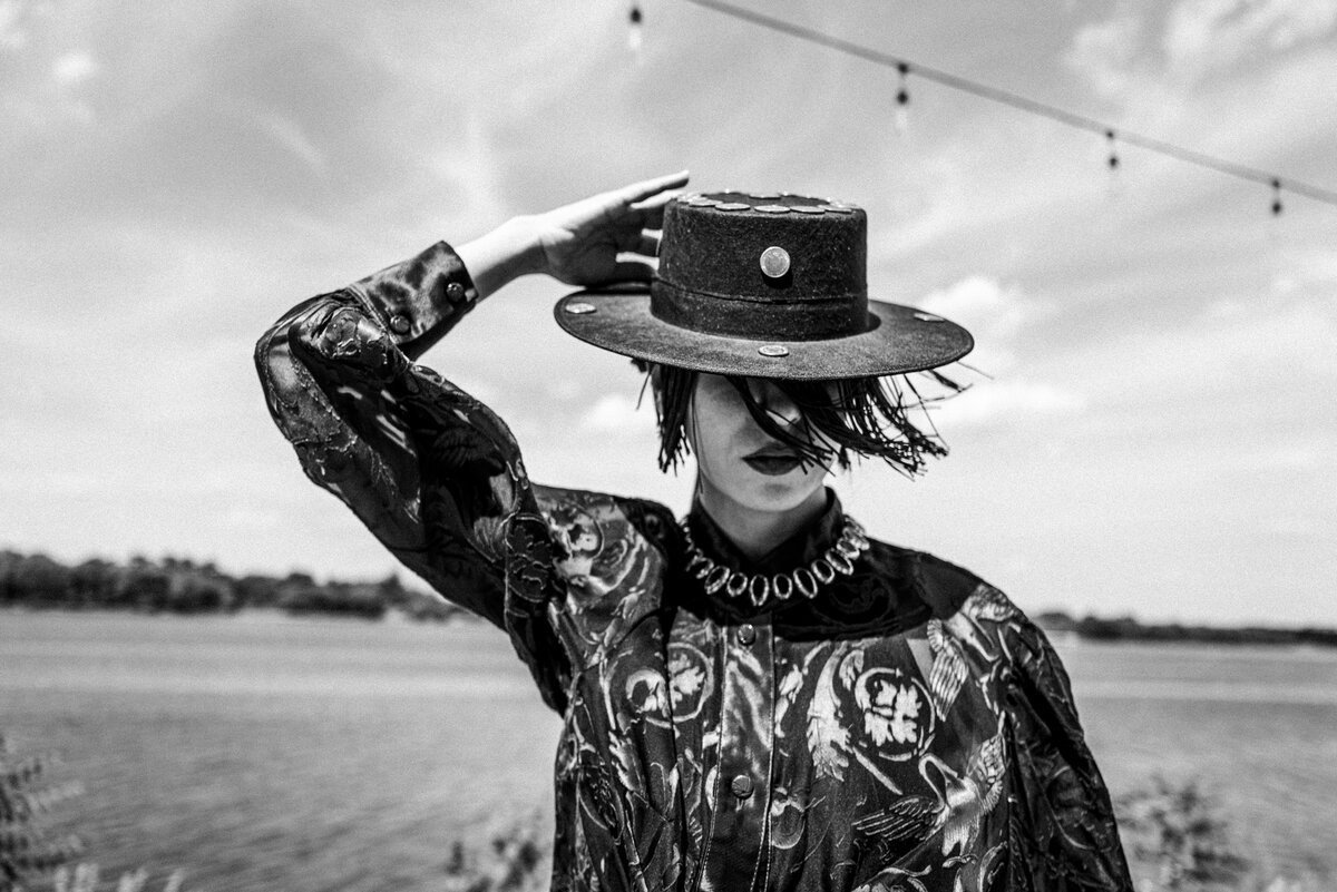 styled woman in printed jacket and wide-brim hat shading face, lakeside