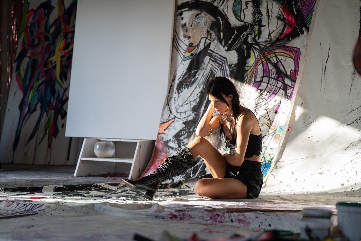 woman seated in a paint-splattered artist's studio against graffiti canvases