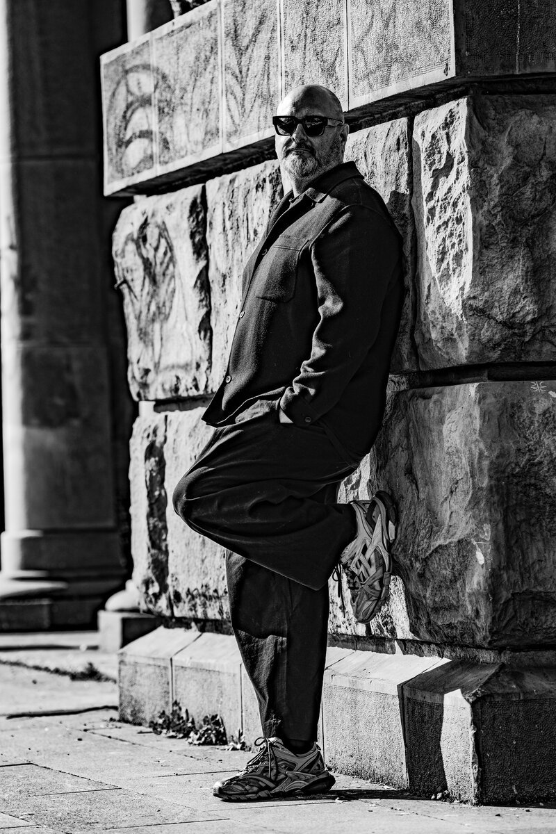 bearded bald man in sunglasses and long coat leaning against stone building