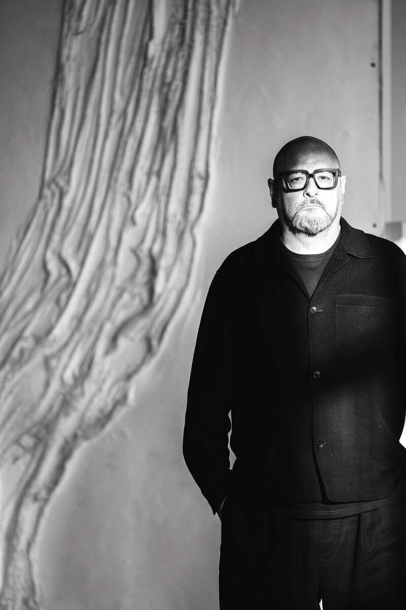bald bearded man in glasses, black shirt, against textured wall