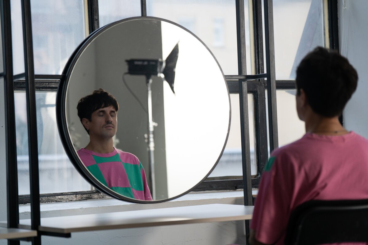 man in pink/green colorblock tee, eyes closed, reflected in a round mirror by a studio window