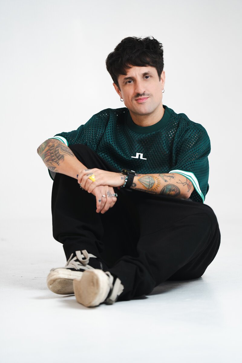 tattooed man with mustache seated on floor, green mesh tee, against white seamless