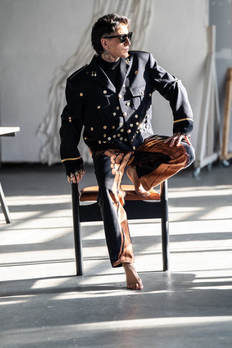 man in gold-buttoned military-style jacket and printed trousers seated on chair, sunlit loft