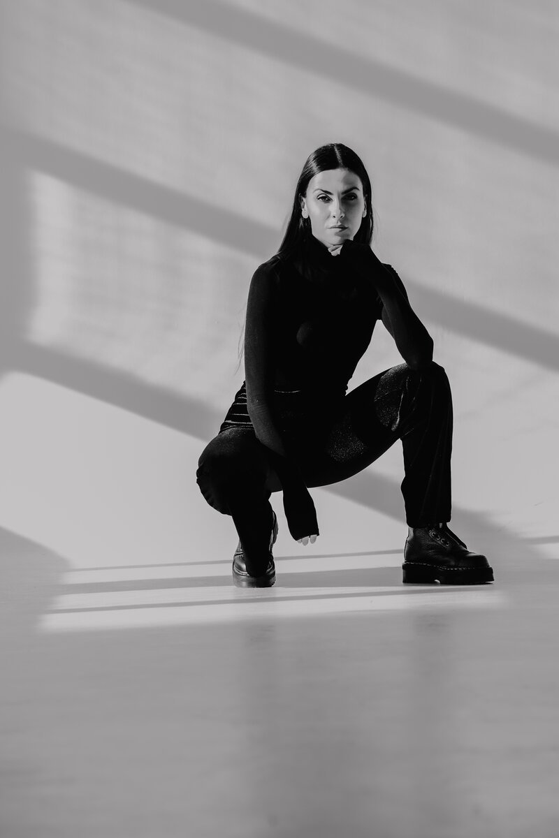 woman in black turtleneck and trousers crouching in a bright window-lit room, B&W