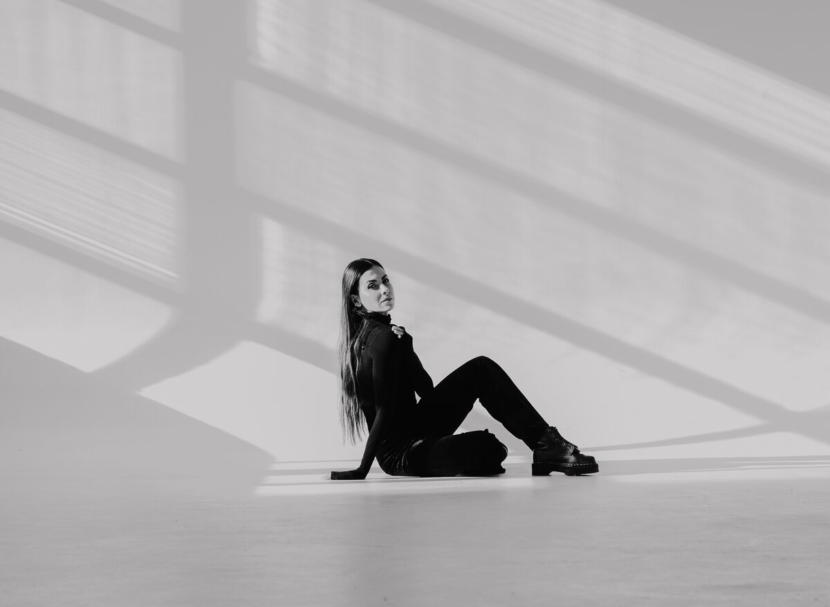 woman in all-black outfit and boots seated on floor of bright, minimal light-drenched space