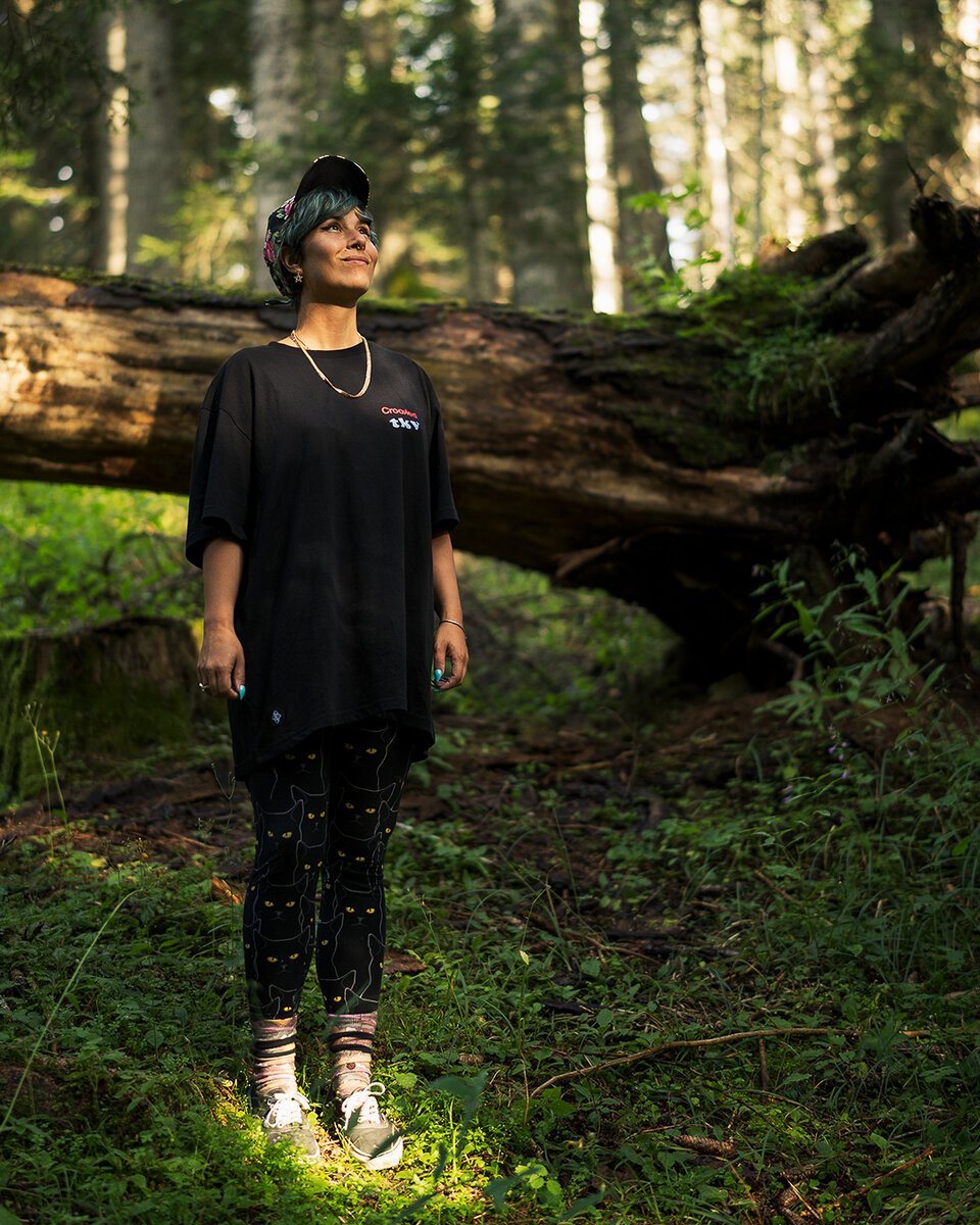 woman in oversized black Crooked tee, cat-print leggings and cap, standing in a sunlit forest by a fallen log